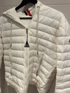 Vit dunjacka från Moncler med huva - Snygg vit dunjacka från Moncler med quiltad design, dragkedja framtill och två sidofickor med dragkedja. Jackan har huva med röd, blå och vit randig detalj samt justerbara snören. Perfekt för kyliga dagar och riktigt stilren look.