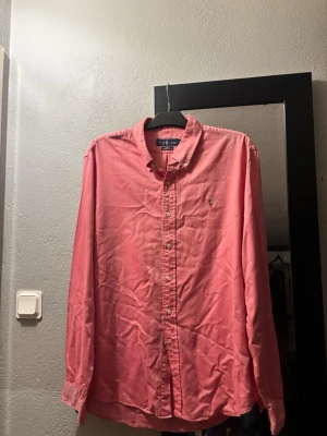 Rosa skjorta från Ralph Lauren - Klassisk rosa skjorta från Ralph Lauren med button-down krage och broderad logga på bröstet. Skjortan har långa ärmar och knäppning framtill. Perfekt för en stilren och avslappnad look.