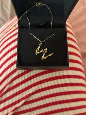 Guldhalsband med färgglada stenar - Snyggt halsband i guld med hängsmycke format som bokstaven w, dekorerat med kristaller. Vet tyvärr inte vart smycket är från då det var en present💞