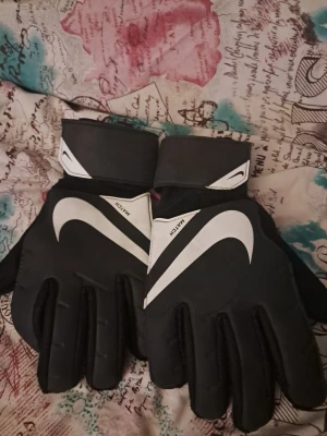 Svarta Nike målvaktshandskar - Säljer ett par svarta målvaktshandskar från Nike med vit logga och breda kardborreband vid handleden. Handskarna har en sportig design och är perfekta för fotbollsmatcher eller träning. Materialet är syntetiskt och greppvänligt.