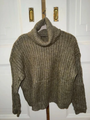 Olivgrön stickad polotröja - Mysig olivgrön stickad polotröja med grov ribbstickning och oversized passform. Tröjan har långa ärmar och en hög, bred polokrage som ger extra värme. Perfekt för kalla dagar och enkel att styla till jeans eller kjol.