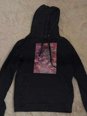 Svart hoodie med konstmotiv🩷 - Svart hoodie med stor huva och dragsnören. På bröstet finns ett färgstarkt konstmotiv i rosa och vitt som föreställer ett ansikte. Hoodien har en klassisk känguruficka framtill och ribbade muddar. Perfekt för dig som gillar streetwear eller Stockholmsstil, och unika prints🖤🩷