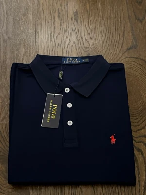 Mörkblå pikétröja från Ralph Lauren - Klassisk mörkblå pikétröja från Ralph Lauren med röd broderad logga på bröstet. Tröjan har krage, tre vita knappar framtill och är tillverkad i mjuk bomull. Perfekt för en clean och stilren look.