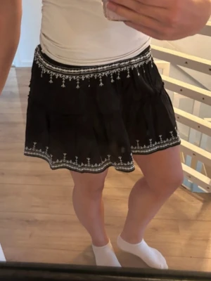 Svart kjol med broderi - Supersöt svart kortkjol med vita broderade detaljer längs midjan och nederkanten. Kjolen har en luftig passform och elastisk midja som gör den bekväm att bära. Perfekt till sommaren och enkel att matcha med olika toppar. Från hm storlek xxs men passar även xs. Lätt genomskinlig men inte mycket. Aldrig använd