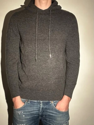 Mörkgrå stickad hoodie - Mysig mörkgrå stickad hoodie med huva och dragsko. Tröjan har en enkel och stilren design utan tryck eller logga, och är tillverkad i ett mjukt material som passar perfekt för avslappnade dagar. Långa ärmar och ribbade muddar ger en skön passform.