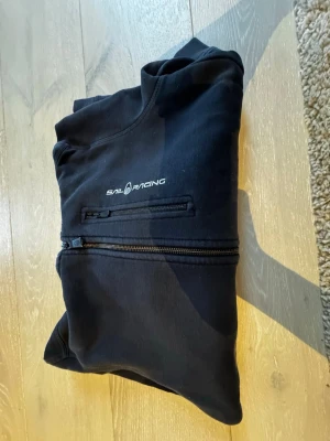 Svart hoodie från Sail Racing - Säljer en svart hoodie från Sail Racing med dragkedja över bröstet och en mindre ficka med dragkedja. Hoodien har en klassisk huva och är tillverkad i mjukt material, perfekt för en avslappnad stil. Diskret logga på bröstet.