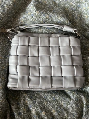 Ljusgrå quiltad axelväska i skinn - Trendig ljusblå handväska med flätad design och mjukt skinnmaterial. Väskan har både ett kort handtag och en avtagbar axelrem för olika stilar. Perfekt accessoar för dig som gillar stilrena detaljer och modern look. har en lite fläck på väskan men syns knappt!