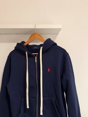 Marinblå hoodie från Polo Ralph Lauren - Snygg marinblå hoodie från Polo Ralph Lauren med dragkedja, vit dragsko och klassiska röda loggan broderad på bröstet. Tillverkad i mjuk bomullsblandning och har två fickor framtill samt en skön huva. Helt ny och oanvänd. Lappen finns kvar på.