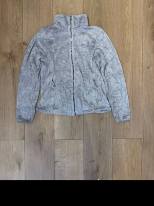 Ljusgrå teddyjacka från The North Face - Supermjuk och fluffig teddyjacka från The North Face i ljusgrå färg. Jackan har hög krage, dragkedja framtill och två sidofickor. Perfekt för att hålla värmen och samtidigt se stilren ut.