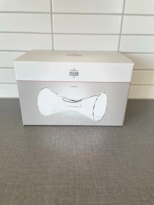 Holmegaard Urania trådlös högtalare - Stilren trådlös högtalare från Holmegaard i modellen Urania. Högtalaren har en unik, böjd form och är tillverkad i transparent glas som ger en modern känsla. Perfekt för dig som vill ha snygg design och bra ljud i ett. Levereras i originalförpackning.