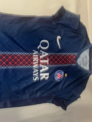 PSG matchtröja blå Nike - Snygg blå PSG matchtröja från Nike med Qatar Airways-tryck på bröstet och klubbmärke med stjärna. Tröjan har korta ärmar och rund hals, samt ett rött och vitt mönster längs mitten. Perfekt för fotbollsfans som vill representera sitt lag.