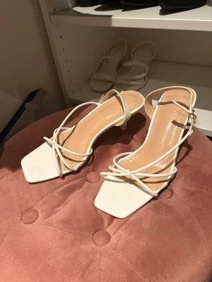 Vita klackar  - Säljer ett par vita klackar  med smala remmar och fyrkantig tå. Skorna har låg klack och spänne vid ankeln. Tillverkade i syntetmaterial och perfekta för sommarens alla outfits. Aldrig använda. 