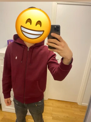 Vinröd hoodie från Polo Ralph Lauren - Snygg vinröd hoodie från Polo Ralph Lauren med dragkedja, huva med dragsko och två fickor framtill. Ikoniska vita loggan broderad på bröstet. Perfekt för en chill och stilren look. Tillverkad i mjukt bomullsmaterial.
