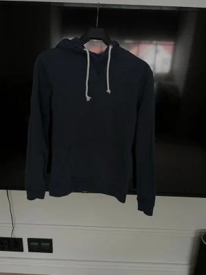 Mörkblå hoodie från Pier One - Mörkblå hoodie från Pier One med klassisk känguruficka och vita dragsnören i huvan. Hoodien har en avslappnad passform och är tillverkad i mjukt material, perfekt för en chill och stilren look.