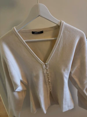 Beige kofta med knappar Gina Tricot - Snygg beige kofta från Gina Tricot med v-ringning och dekorativa tygklädda knappar framtill. Koftan har lång ärm och är tillverkad i ett mjukt, stretchigt material som känns skönt mot huden. Perfekt att styla till jeans eller kjol.