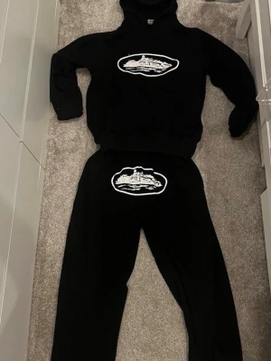 C0rteiz tracksuit - Säljer ett svart loungewear-set med matchande hoodie och byxor. Båda plaggen har ett vitt tryck med motiv av en färja eller båt på bröstet och höften. Setet är i mjukt material och har en avslappnad passform, perfekt för chill dagar hemma.