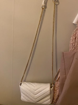 Vit quiltad axelväska med guldkedja - Snygg vit axelväska med quiltad design och guldfärgad kedjerem. Väskan har en stilren och modern look, perfekt för att lyfta vilken outfit som helst. Tillverkad i syntetmaterial och har en kompakt, rektangulär form.