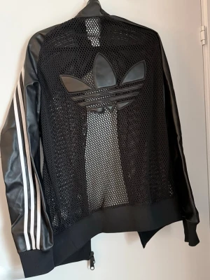 Svart mesh bomberjacka Adidas - Unik svart bomberjacka från Adidas med mesh-tyg på fram- och baksida, stora Trefoil-loggan bak och klassiska vita ränder på ärmarna. Jackan har dragkedja framtill och ribbade muddar. Perfekt för en sportig och trendig streetstyle. Enbart använd två tillfällen, nyskick. Köptes för; 1345 kr sommar 2025.