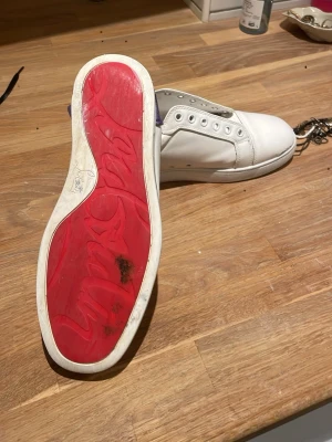 Vita sneakers från Christian Louboutin - Säljer ett par vita sneakers från Christian Louboutin med klassisk röd sula och snörning. Skorna är tillverkade i skinn och har en stilren, rund tå samt silverfärgade öljetter. Perfekta för dig som vill ha något exklusivt och trendigt.pris kan sänkas vid snabb affär 