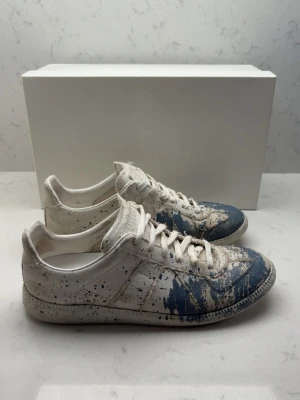 Maison Margiela blå-vita sneakers - Unika sneakers från Maison Margiela i vitt skinn med blå färgstänk och konstnärlig finish. Lågt skaft, rund tå och klassisk snörning. Snygg kontrast mellan det vita och blå detaljerna ger en cool streetwear-vibe.