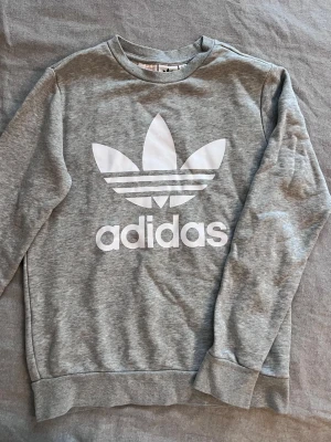 Grå sweatshirt från Adidas - Säljer en grå sweatshirt från Adidas med stort vitt Trefoil-tryck och logga på bröstet. Tröjan har rund halsringning, långa ärmar och ribbade muddar vid ärmslut och nederkant. Perfekt för en avslappnad och sportig stil.