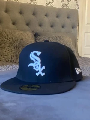 Svart Chicago White Sox keps New Era - Snygg svart keps från New Era med Chicago White Sox-logga broderad i vitt framtill. Klassisk flat brim och MLB-logga baktill. Perfekt för dig som gillar streetwear och sportiga accessoarer.