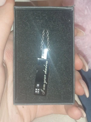 Silvrigt halsband med text och hjärta - Snyggt halsband i silverfärgad metall med rektangulärt hänge. Hänget har texten 'I am your shadow' ingraverat på framsidan och tre små glittriga stenar. Baksidan har ett litet vitt hjärta. Perfekt accessoar för en personlig touch.