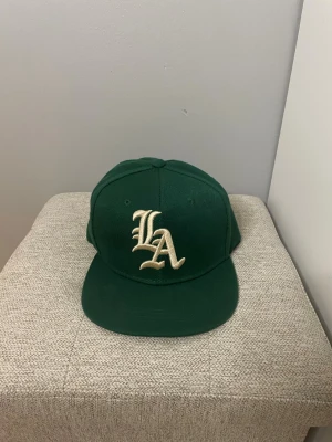 Grön LA keps med broderad logga - Snygg grön keps med stor broderad LA-logga i vitt framtill. Klassisk rak skärm och justerbar baksida. 