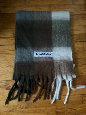 Rutig halsduk från Acne Studios - Mjuk och fluffig halsduk från Acne Studios i rutigt mönster med jordnära toner av brunt, beige, grått och vitt. Halsduken har långa fransar i ändarna och en tydlig Acne Studios-logga. Perfekt för att addera stil till din outfit.