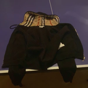 Svart Burberry hoodie med dragkedja - Svart hoodie från Burberry med klassiskt rutigt foder i huvan. Tröjan har dragkedja framtill, snörning i huvan och ribbade muddar. Perfekt för dig som vill ha en stilren och lyxig streetwear-look. Nästan inte använd alls. storlek M men känns som S
