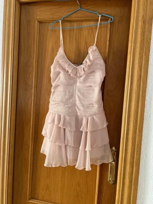 Ljusrosa volangklänning med smala axelband - Söt ljusrosa kortklänning med smala axelband och volangdetaljer både vid urringningen och kjolen. Klänningen har en omlottkänsla och är tillverkad i ett lätt, skirt material som ger ett romantiskt intryck.