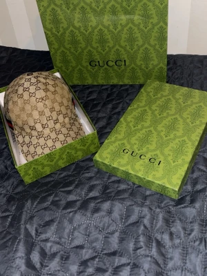 Gucci beige keps med GG-mönster - Snygg beige keps från Gucci med klassiskt GG-monogram över hela kepsen och en grön-röd rand på sidan. Kepsen har böjd skärm och justerbar rem bak. Tillverkad i canvasmaterial med exklusiv känsla. Perfekt för dig som vill ha en trendig accessoar med lyxig touch.
