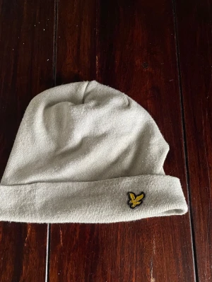 Ljusgrå mössa från Lyle & Scott - Snygg ljusgrå mössa från Lyle & Scott med klassisk gul broderad örnlogga framtill. Mjuk och stretchig stickad modell som passar perfekt till kalla dagar. Enkel design som funkar till det mesta.