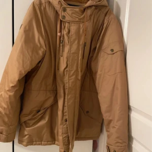 Beige tjockvadderad jack med huva, herr storlek 2XL - Snygg beige tjockvadderad  med huva och dragkedja framtill. Jackan har flera fickor med knappar och dragkedjor samt snörning i halsen för extra stil. Perfekt för kalla dagar och enkel att matcha med olika outfits.