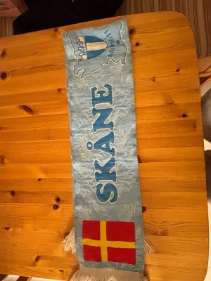 Ljusblå Malmö FF Skåne-halsduk retro från 2011 - Säljer en ljusblå halsduk med Malmö FF och Skåne-tryck. Ena sidan har Skånes flagga i rött och gult, andra sidan har svensk flagga i blått och vitt. Tydliga broderade detaljer och fransar i ändarna. Perfekt för dig som vill visa ditt stöd för MFF och Skåne.