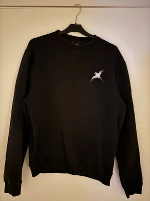 Svart sweatshirt från Axel Arigato - En svart sweatshirt med rund hals och ett litet vitt fågeltryck på bröstet. Tröjan har långa ärmar och ribbade muddar vid ärmslut och nederkant. Mycket bra skick, nypris ca 1199.