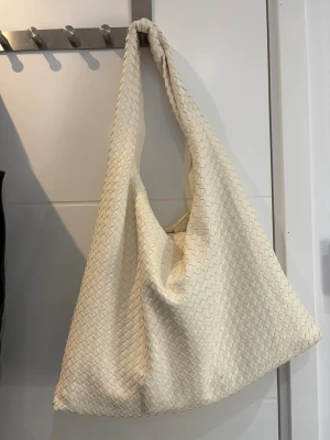 Beige axelväska i flätad skinnimitation - Stilren beige axelväska med flätad design och mjuk, slouchy form. Väskan har bred axelrem och rymmer allt du behöver till vardagen. Perfekt för dig som gillar minimalistisk och trendig look. Lite smink och smuts på insidan men helt orörd utsida