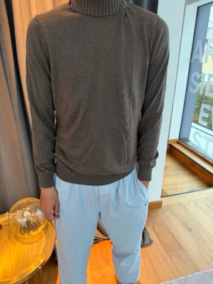 Brun polotröja i mjukt material - Mysig brun polotröja med ribbad krage och långa ärmar. Tröjan har en enkel och stilren design, perfekt för att chilla hemma eller när du vill ha något bekvämt. Materialet ser ut att vara mjukt och stretchigt, vilket gör den extra skön att bära.