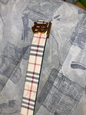 Rutigt bälte med metallspänne - Snyggt bälte med klassiskt rutigt mönster i beige, svart och rött. Bältet har ett coolt metallspänne i guld med unik design. Perfekt accessoar för att lyfta din outfit och ge en trendig touch. Materialet känns slitstarkt och mönstret är riktigt stilrent.