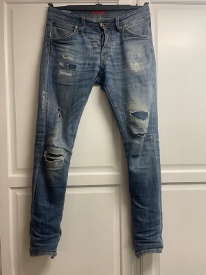 Blå slitna jeans med hål - Säljer ett par blå jeans med slitningar och hål på både fram- och baksida. Jeansen har klassisk femficksmodell, normal passform och är tillverkade i bomull. Perfekta för en avslappnad och trendig look.