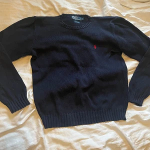 Mörkblå stickad tröja från Polo Ralph Lauren - Klassisk mörkblå stickad tröja från Polo Ralph Lauren med rund halsringning och den ikoniska röda logotypen broderad på bröstet. Tröjan har ribbade muddar vid ärmslut och nederkant. Perfekt för lager på lager och enkel att matcha.