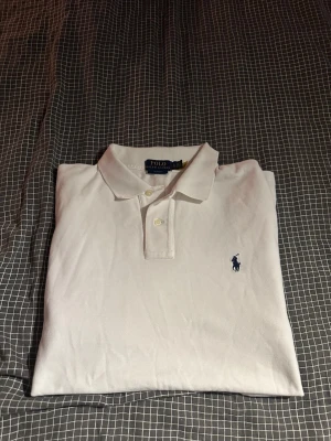 Vit pikétröja från Polo Ralph Lauren - Klassisk vit pikétröja från Polo Ralph Lauren med blå broderad logga på bröstet. Tröjan har korta ärmar, krage och knappar framtill. Tillverkad i mjuk bomull som känns skön mot huden. Perfekt till jeans eller shorts för en clean look.