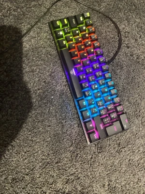 RGB mekaniskt tangentbord med sladd - Säljer ett kompakt mekaniskt tangentbord med RGB-belysning. Tangentbordet är svart med färgglada bakgrundsljus som kan justeras. Ansluts via USB-kabel och har en minimalistisk design utan numerisk del. Perfekt för gaming eller skrivbordet.