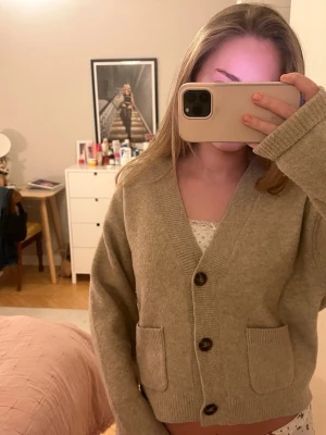 Stickad kofta  - Mysig beige kofta från Uniqlo i storlek XS. Koftan har v-ringning, stora knappar framtill och två fickor. Tillverkad i ett mjukt material som känns skönt mot huden. Perfekt att slänga på sig över en topp.