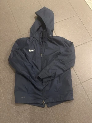 Mörkblå Nike Shield vindjacka 147-158 (L barn) - Nike Shield vindjacka i mörkblått med huva och dragkedja framtill. Jackan har meshfoder på insidan och klassisk Nike-logga på bröstet. Perfekt för blåsiga dagar och enkel att matcha med det mesta.