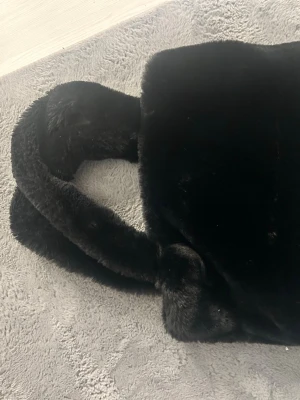 Svart fluffig handväska i fuskpäls - Mysig svart handväska i fluffig fuskpäls med två rejäla handtag. Väskan har ett innerfack med dragkedja och är helsvart både på utsidan och insidan. Perfekt accessoar för dig som gillar mjuka material och vill sticka ut lite extra.