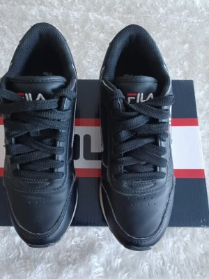 Svarta sneakers från Fila - Stilrena svarta sneakers från Fila Orbit med klassisk design. Skorna har rund tå, snörning och vit sula som kontrast. Tillverkade i syntetmaterial med diskreta detaljer och Fila-logga på plösen. Perfekta för en sportig och avslappnad look. Använda fåtal gånger. Orginalkartong: 31×21×12.