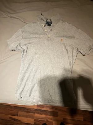 Grå pikétröja från Polo Ralph Lauren - Riktigt fet grå pikétröja från Polo Ralph Lauren med korta ärmar och knappslå framtill. Tröjan har den ikoniska broderade loggan på bröstet och är tillverkad i mjuk bomull för en bekväm känsla. Pris kan diskuteras 