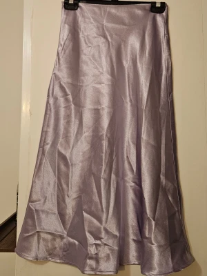 Ljuslila satinkjol - Snygg ljuslila långkjol i satin med glansig finish och lätt utsvängd form. Perfekt för dig som vill ha en stilren och trendig look. Kjolen har en enkel design utan mönster eller detaljer och faller snyggt längs benen. Ser ostruken ut på bilden.