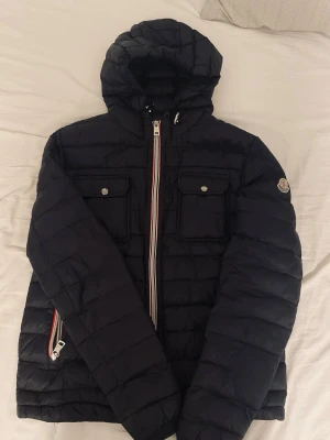 Svart Moncler dunjacka med huva - Snygg svart dunjacka från Moncler med huva och två bröstfickor med knappar. Jackan har quiltad design, dragkedja med Monclers signaturband och logga på ärmen. Perfekt vår/höst jacka.